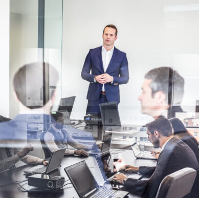 Successful team leader and business owner leading informal in-house business meeting. Business people working on laptops in foreground and glass reflections. Business and entrepreneurship concept.