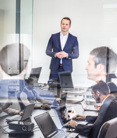 Successful team leader and business owner leading informal in-house business meeting. Business people working on laptops in foreground and glass reflections. Business and entrepreneurship concept.